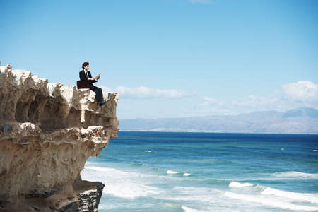 Breaking free from the rat-race. Young businessman sitting on the edge of a cliff overlooking the...