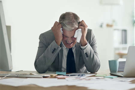 This paperwork is driving me crazy. Shot of a frustrated businessman crumpling up paperwork while sitting at his desk.の写真素材