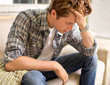 This headache is killing me. A young man sitting on a couch with his hand in his hair as if in pain.