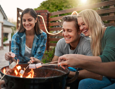 I love roasting marshmallows around a fire. Shot of a group cheerful young friends holding up marshmallows on sticks over a fire outside.の写真素材