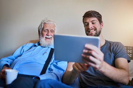 Welcoming grandpa to the age of technology. Shot of a young man showing his elderly grandfather how to use a tablet while sitting on the couch at home.の写真素材