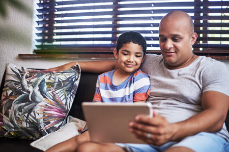 Next episode. Cropped shot of a cheerful young man and his son watching videos on a digital tablet together while hanging out on a sofa at home during the day.の写真素材