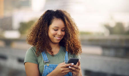 This is such a funny text. Cropped shot of an attractive young woman standing outdoors and looking down while using her cellphone.の写真素材