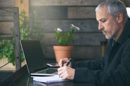 Making some notes. Shot of a mature businessman writing notes while using his laptop in a cafe.の写真素材