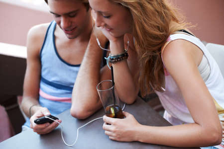 Get closer to your crush through the wonders of technology. Happy teen couple sharing an MP3 player and listening to music while enjoying their beverages.の写真素材