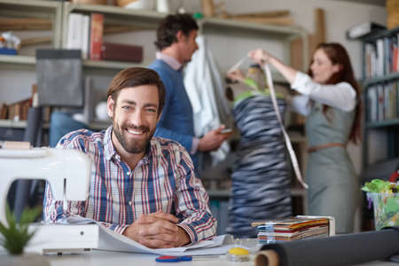We can make anything. Portrait of a young fashion designer sitting in the workshop while his colleagues work in the background.の写真素材