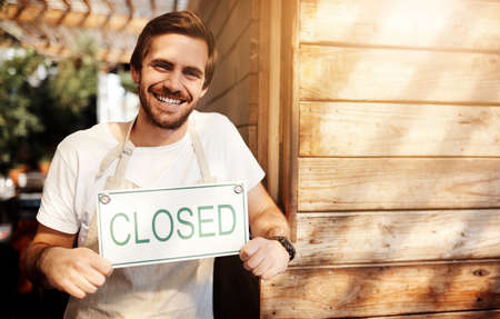 Sorry, were closed. Cropped portrait of a handsome young male business owner holding up a sign in the doorway of his coffee shop.の写真素材