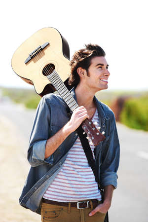 He is a true muso. Smiling young man holding his guitar over his shoulder standing on the side of the road - closeup.の写真素材