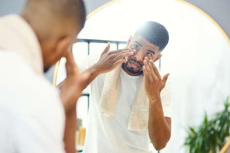 This face cream was well recommended. Young man applying face product in his bathroom mirror.