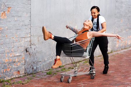 Im about to fly. Full length shot of an energetic young woman pushing her female friend in a shopping cart outdoors.の写真素材