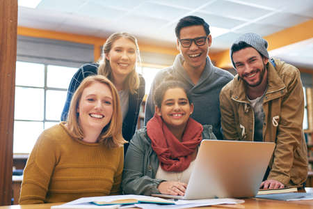 It feels a bit better when youre studying together. Portrait of a cheerful young group of students working together using a laptop to study for exams inside of a library.の写真素材