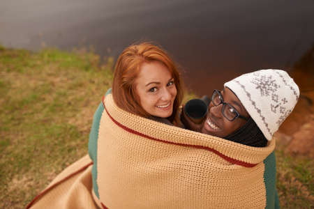 As close as two best friends can get. A high angle portrait of two happy young women huddled under a blanket next to a lake in winter.の写真素材