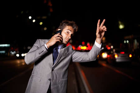 Business never sleeps. A handsome businessman hailing a cab while talking on his cellphone in the city at night.の写真素材