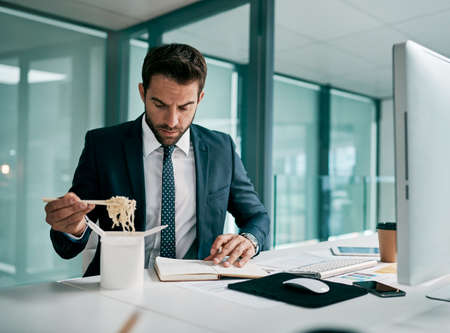 Cant focus on just one thing at a time. Shot of a focused young businessman eating noodles while writing in a notebook inside of the office.の写真素材