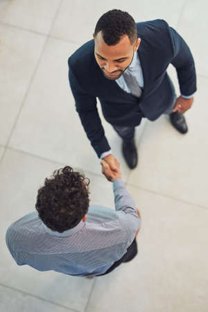 Thanks for stopping by. High angle shot of two confident young businessmen shaking hands in agreement while standing in the office during the day.の写真素材