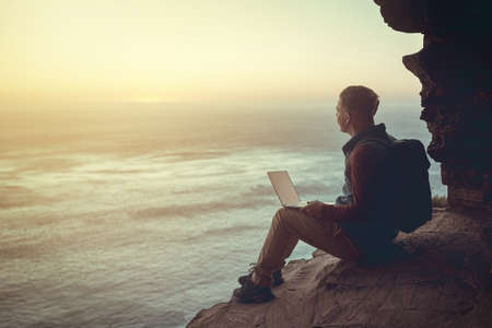 Connecting to the world. Full length shot of a young man using his laptop while sitting on a cliff...