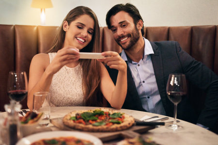 I want to remember this pizza. Shot of a happy young couple taking a picture of their meal on a cellphone during a romantic dinner date at a restaurant.の写真素材