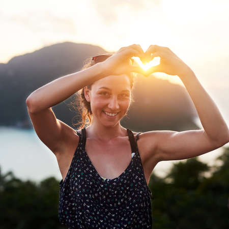 Whats there not to love about summer. Portrait of a young woman making a heart gesture outside.の写真素材