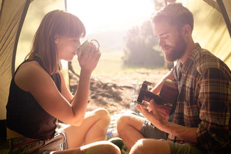 Coffee time at camp. Shot of a young man playing guitar to his girlfriend in a tent.の写真素材