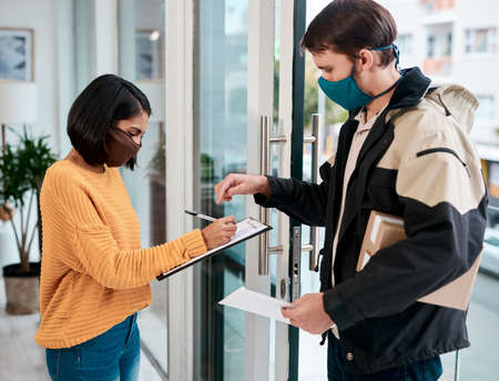 Just your signature and its all yours. Shot of a masked young woman signing for a delivery received at home.の写真素材