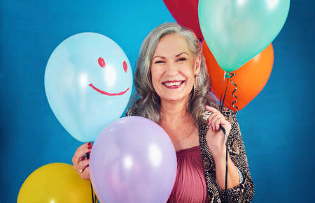 Its my birthday today. Portrait of a cheerful senior woman posing holding balloons in studio against...