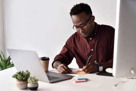 I have to take notes before I forget. Cropped shot of a handsome young businessman sitting alone and making notes while using his laptop in the office.の写真素材
