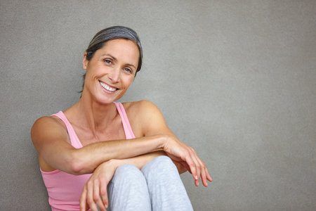 Living a healthy lifestyle. Portrait of an attractive mature woman in gymwear sitting against a gray wall.の写真素材