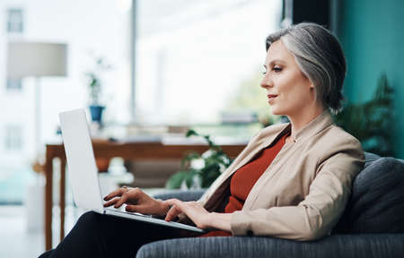 Responding to emails. Cropped shot of an attractive mature businesswoman sitting alone and using a laptop in her home office.の写真素材