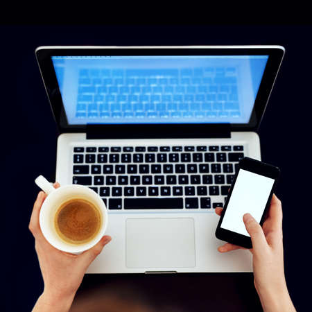 All a blogger really needs. High angle shot of an unidentifiable woman using her phone and laptop while drinking a cup of coffee.の写真素材
