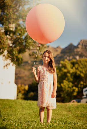 Any bigger and it would carry me away. Portrait of a cute little girl holding a huge balloon while standing outside.の写真素材
