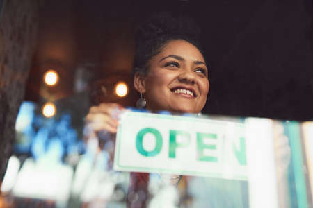 Customers will soon come flooding in. Cropped shot of a young woman hanging up an open sign on the...