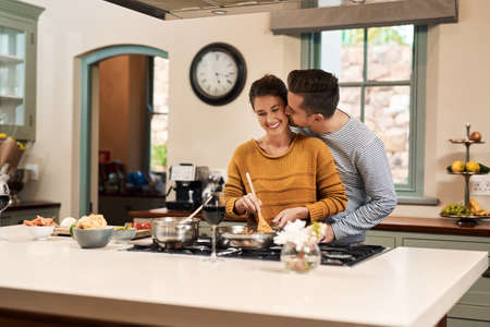 I love you my sweetie pie. Shot of an affectionate young man kissing his wife on the cheek while she prepares a meal in the kitchen at home.の写真素材