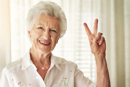 V for vitality. Portrait of a happy elderly woman showing a peace gesture at home.の写真素材