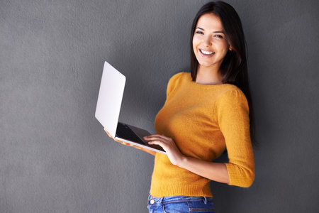 Shes an online social butterfly. Portrait of a beautiful young woman using a laptop against a gray background.の写真素材