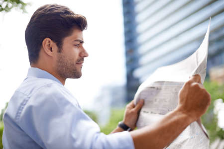 Catching up on the news. A handsome young businessman reading a newspaper while sitting on a bench in the city.の写真素材