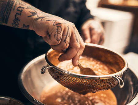 This dish is a knock out. Closeup of a unrecognisable chefs tattooed hands straining food into a bowl in kitchen of a restaurant.の写真素材