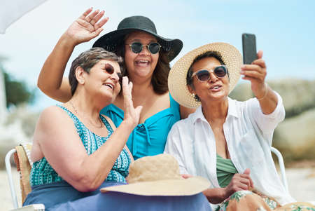 Look at how good Mom looks in a swimsuit. Shot of a mature group of friends using a cellphone for a video chat during a day on the beach.の写真素材
