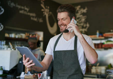 Just calling to let you know your order is ready. Shot of a young barista taking orders via cellphone and logging them on his tablet.の写真素材