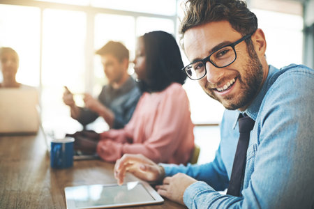 Feeling optimistic about the future of his career. Portrait of a businessman using a digital tablet with his colleagues in the background.の写真素材