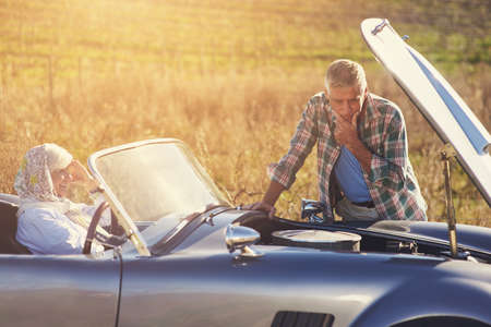 Roadtrip car trouble. Shot of a senior man looking under the hood of his car while on a roadtrip with his wife.の写真素材