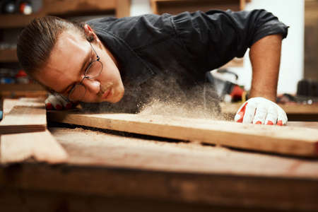 Looking great. Cropped shot of a focused young male carpenter blowing dust off of a piece of wood after sanding it inside of his workshop during the night.の写真素材