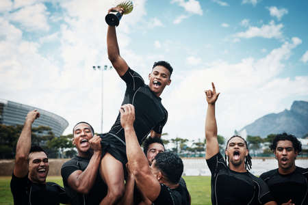 Theyre not just any team theyre rugby cup winners. Cropped shot of a handsome young rugby player holding up a trophy while celebrating with his team on the field.の写真素材