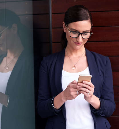 My office in the palm of my hand. Shot of a happy businesswoman using her smartphone next to a window in the office.の写真素材