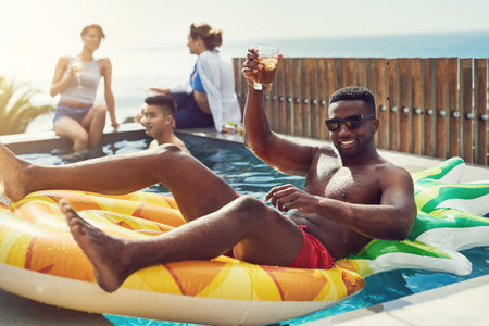The cool guy wishes everyone a happy holiday. Portrait of handsome young man raising up his glass for a toast while relaxing in a pool outdoors with friends.の写真素材