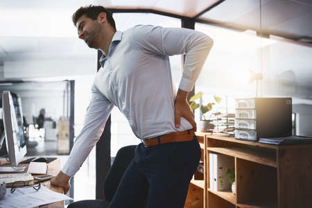 Those cracks dont sound good. Shot of a young businessman suffering with back pain while working late in an office.の写真素材