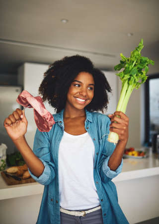 We have all the ingredients well ever need. Shot of a cheerful young woman holding celery in one hand and a piece of meat in the other while standing in the kitchen at home.の写真素材