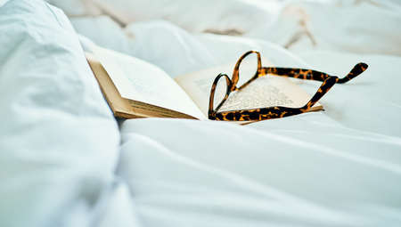 Nothing better than sliding into bed with a good read. Still life shot of a book and a pair of spectacles on a bed.の写真素材