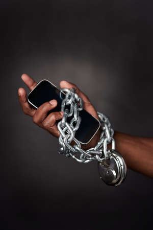 Tied to technology 247. Cropped shot of a mans phone tied to his hand with chains against a black background.の写真素材