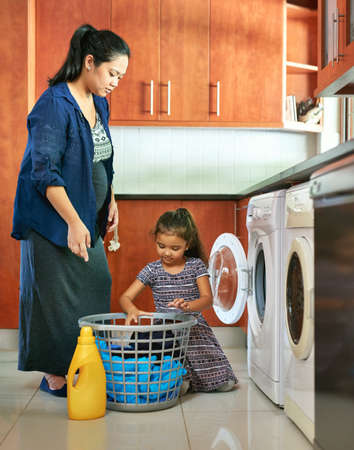Moms little helper. Shot of a pregnant mother and daughter using a washing machine at home.の写真素材