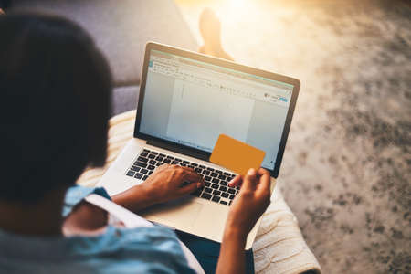 Shopping on her own terms. Shot of a young woman using a laptop and credit card on the sofa at home.の写真素材